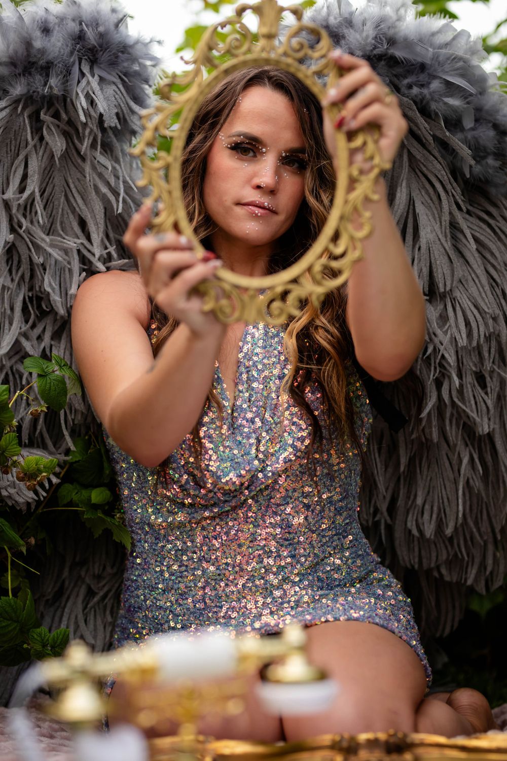 Magical fantasy portrait with gray wings and sequined dress holding ornate gold mirror.