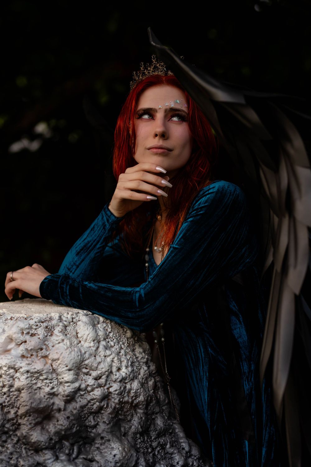 Moody portrait with velvet dress and crown against stone pillar at dusk.