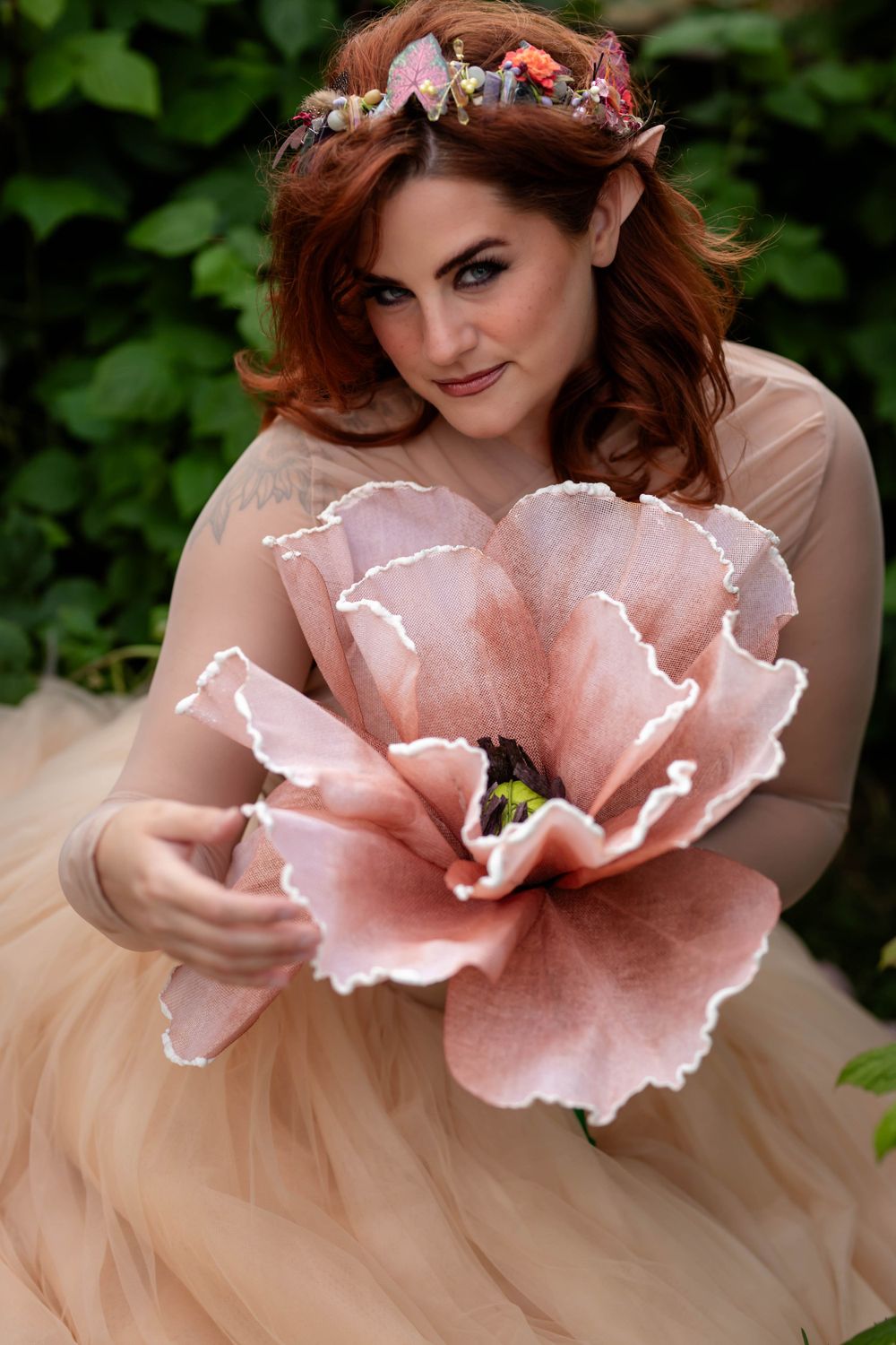 Close-up portrait with floral crown and oversized pink flower against garden backdrop.