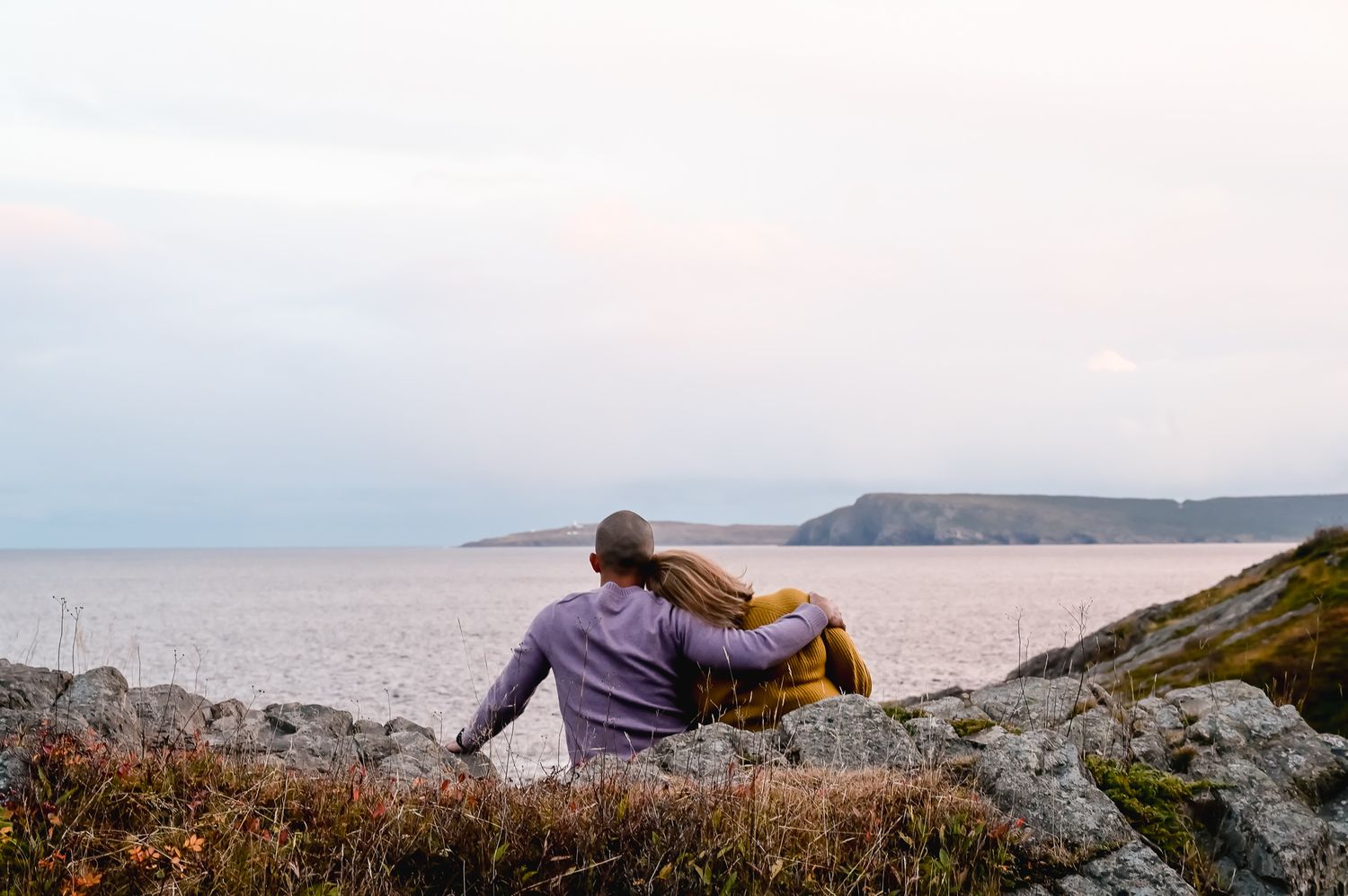 A couple sitting on a rocky shoreline next to the sea at sunset, their silhouettes bathed in warm golden light, St. John's, Newfoundland.