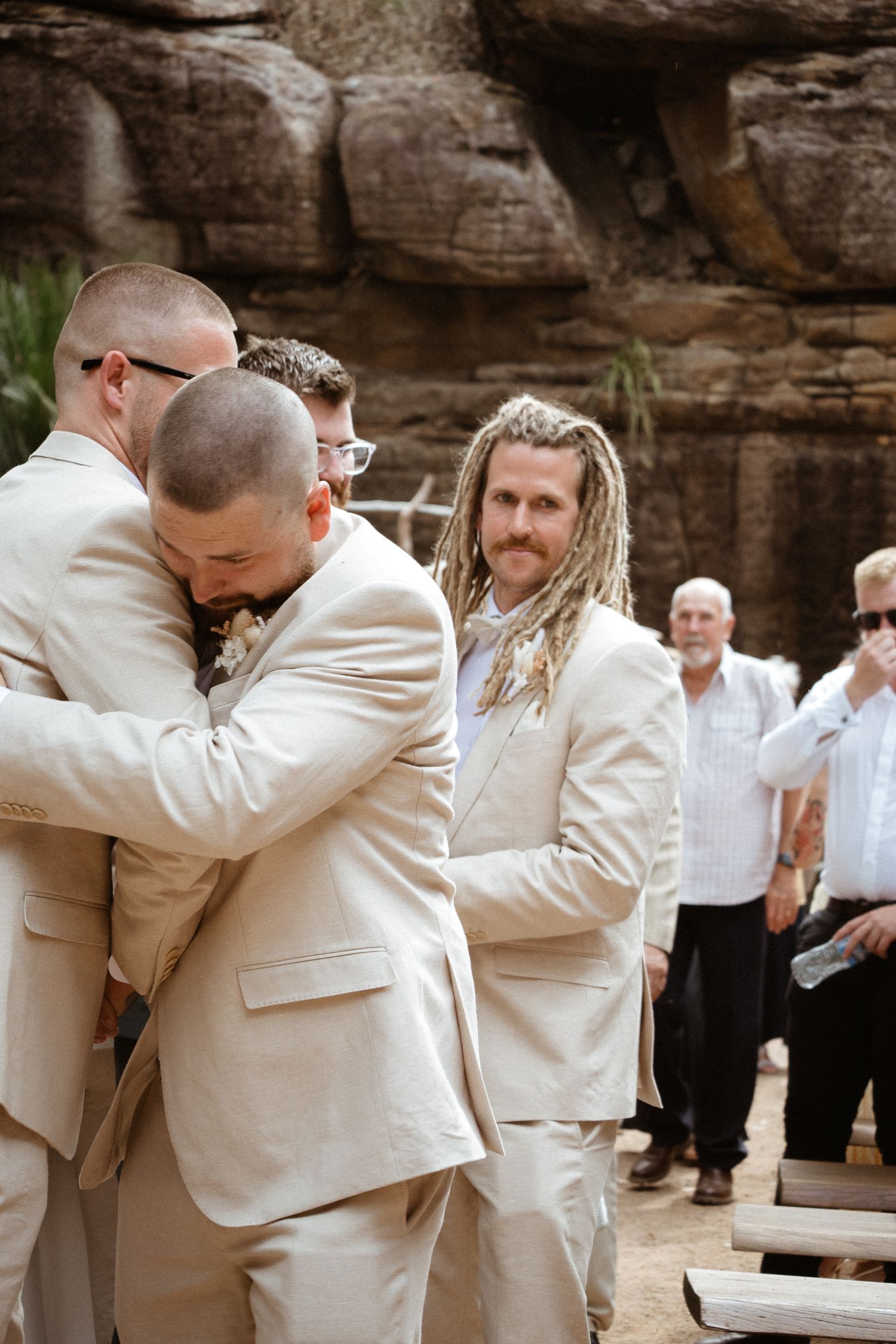 Emotional embrace between wedding attendees in cream-colored suits during outdoor ceremony.