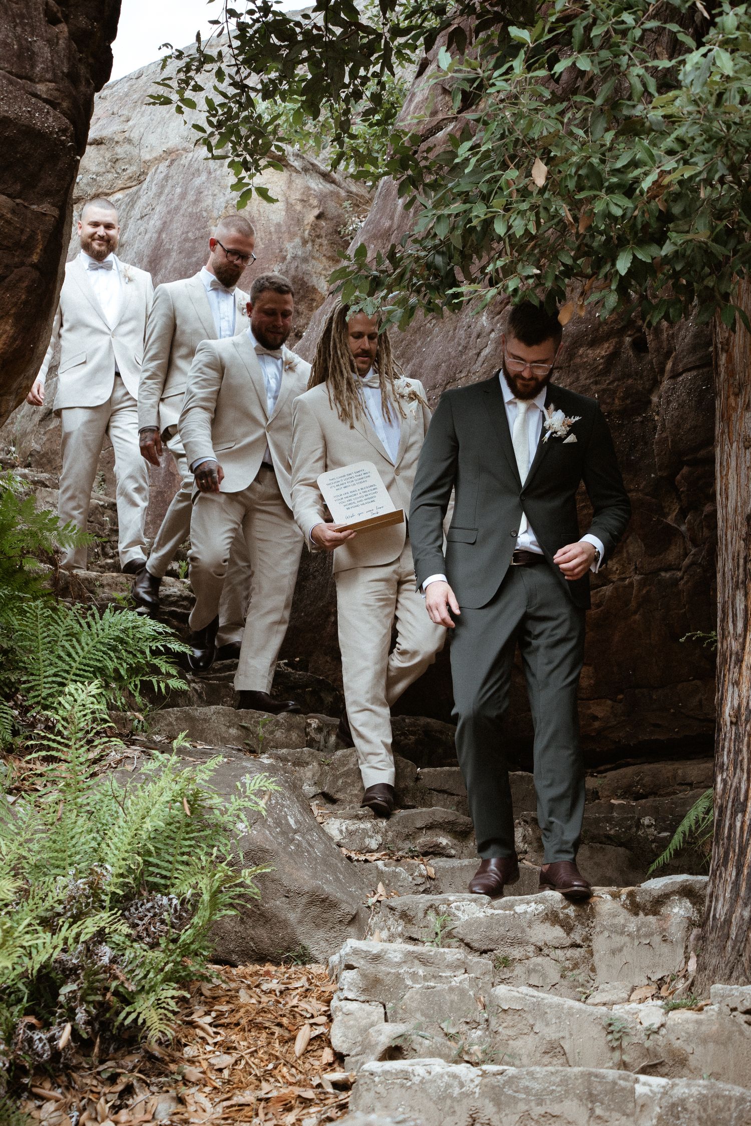 Wedding party descends stone steps in coordinated light suits against a rocky natural backdrop.