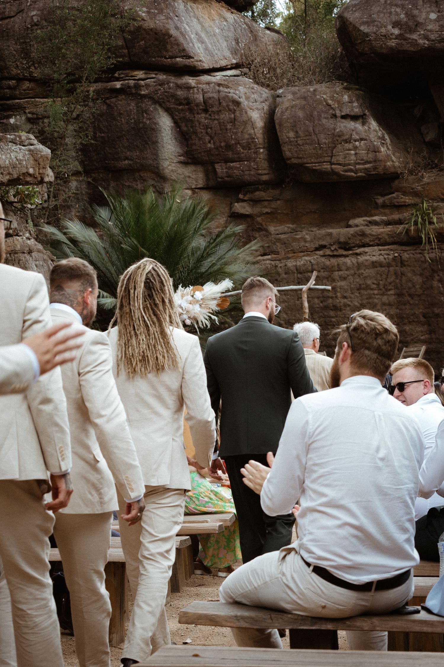 Wedding guests gather near rocky outcrop with ferns during outdoor ceremony.