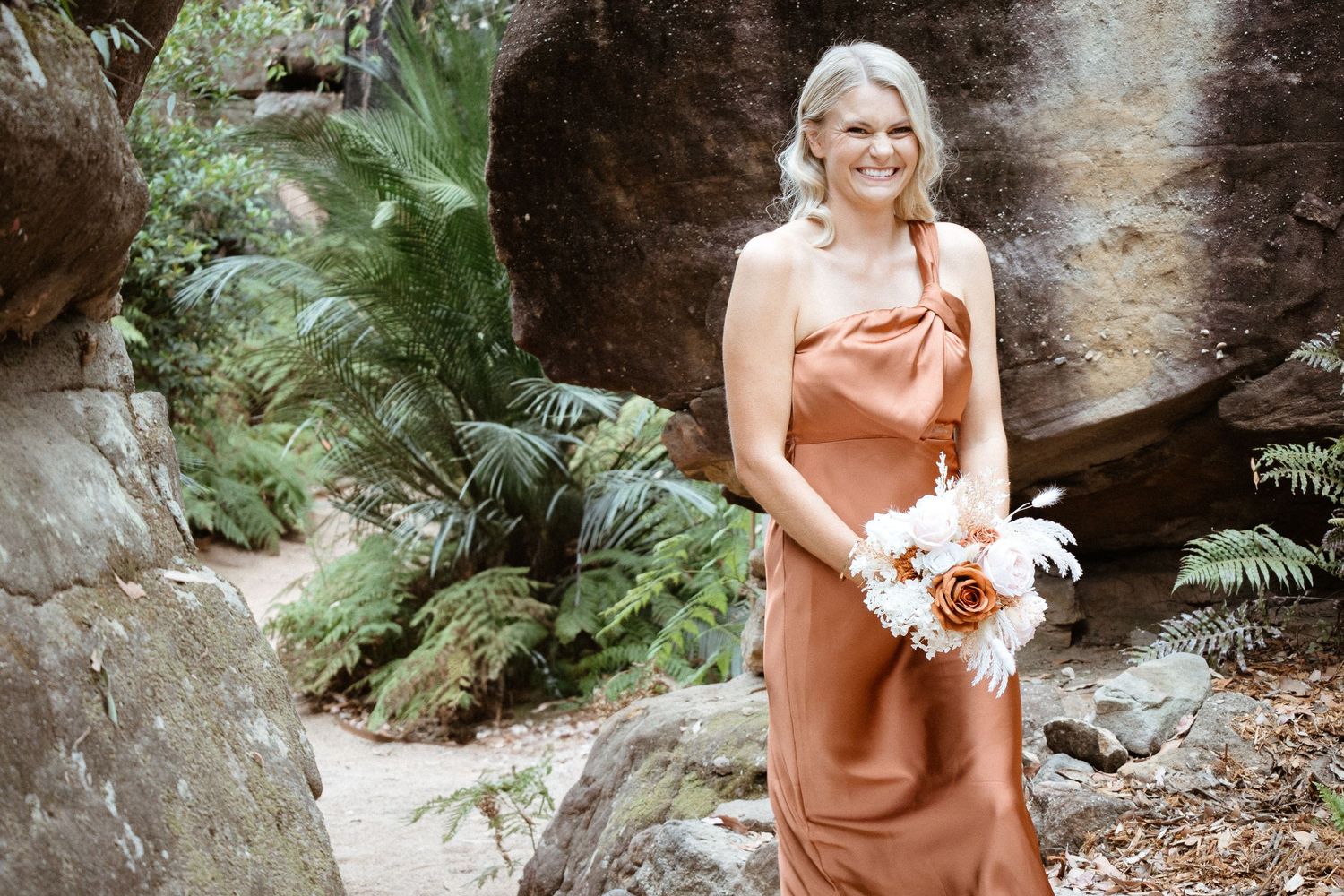 Bridesmaid in copper-colored dress holds white bouquet against natural rock formation.