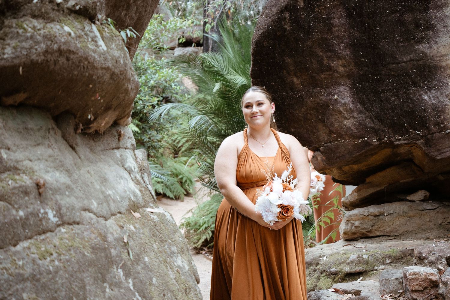 Bridesmaid in rust-colored dress poses with bouquet against rocky landscape with ferns.
