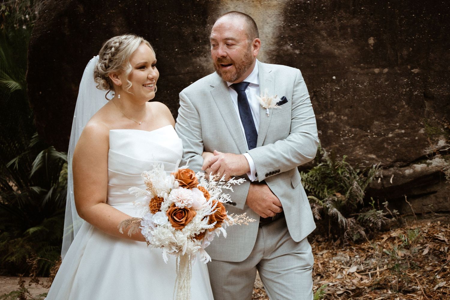 Couple in wedding attire pose with peach and white bouquet against stone backdrop.