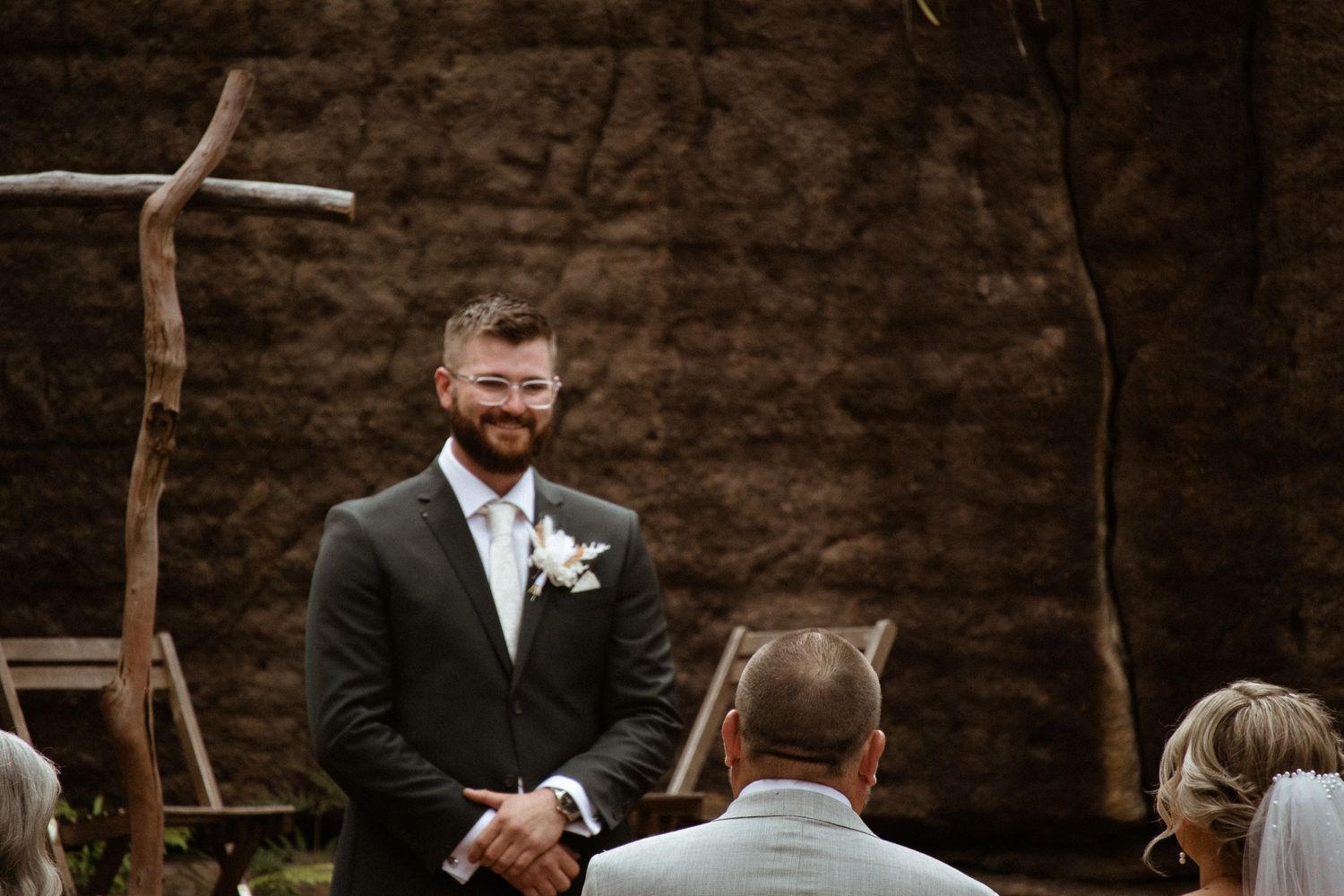 Wedding ceremony takes place in front of rustic wooden cross against stone wall backdrop.