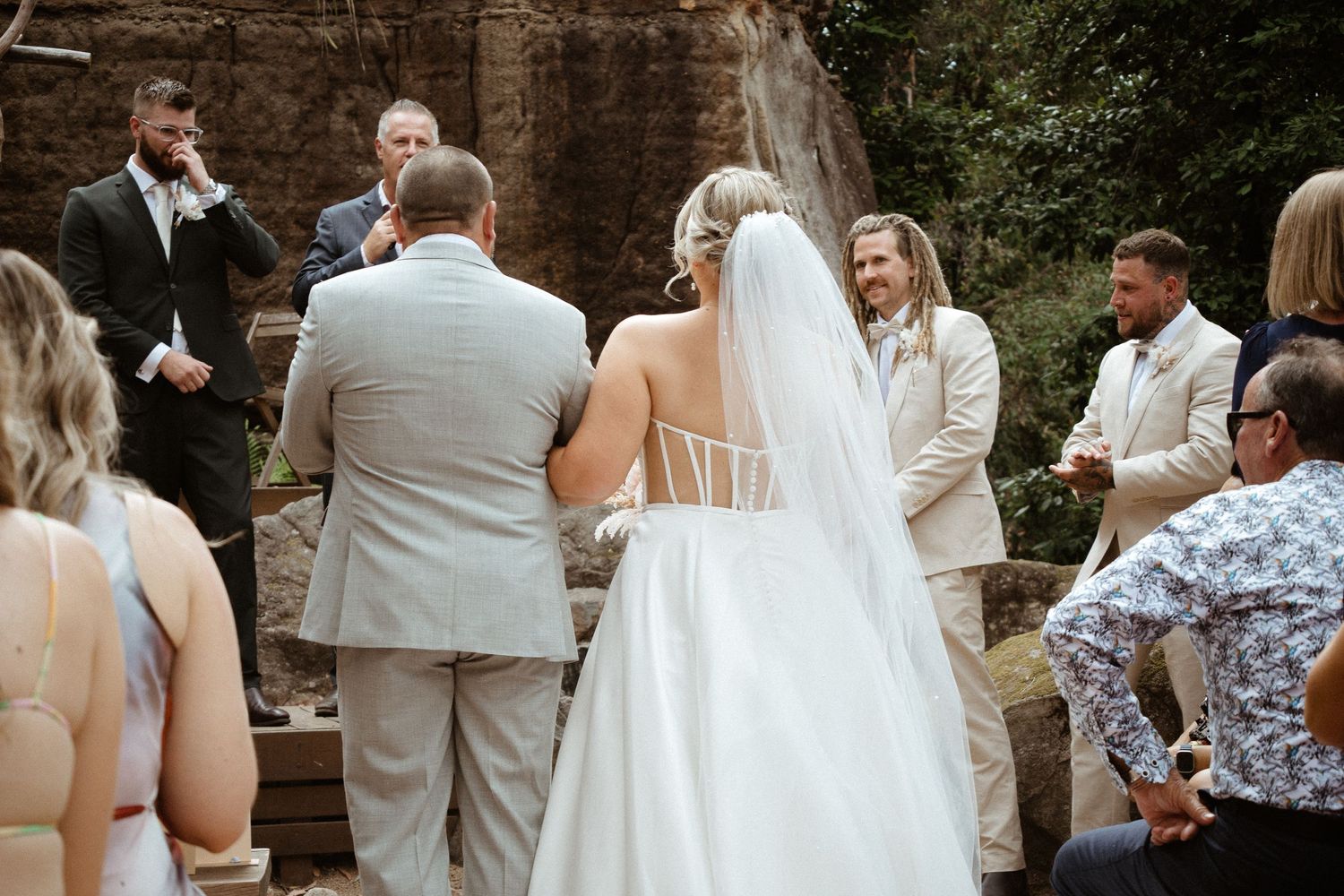 Intimate outdoor wedding ceremony with guests gathered near stone walls and natural elements.