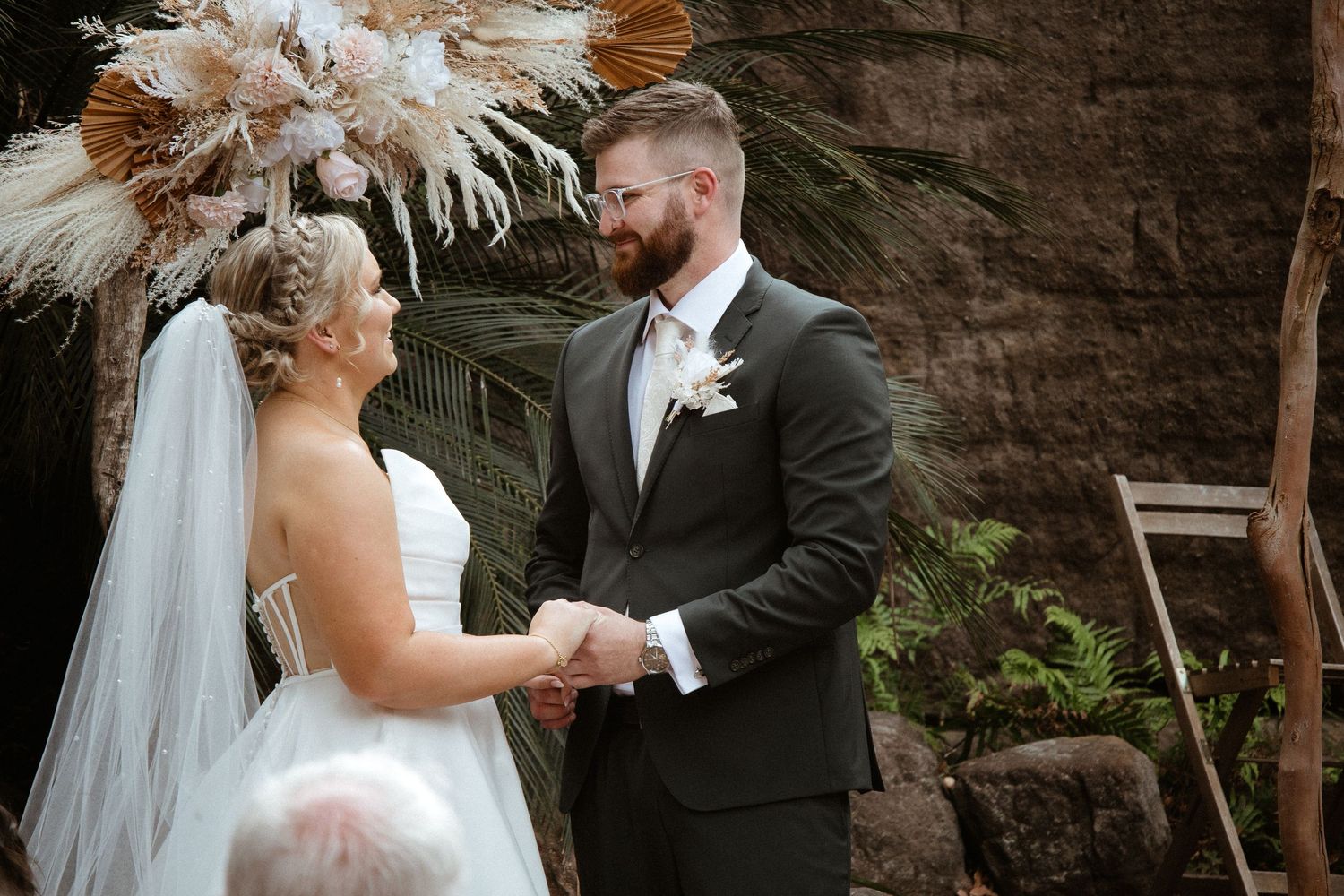 Wedding ceremony under dried palm arrangement against stone backdrop.
