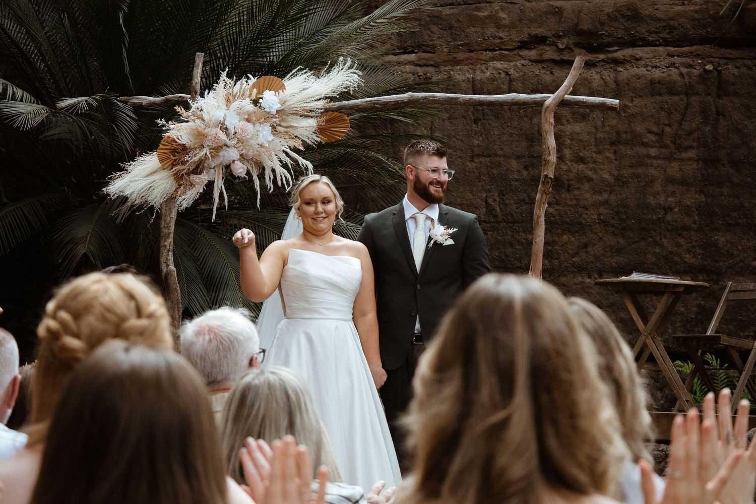 Wedding ceremony takes place under rustic wooden arch with pampas grass decor against stone wall backdrop.
