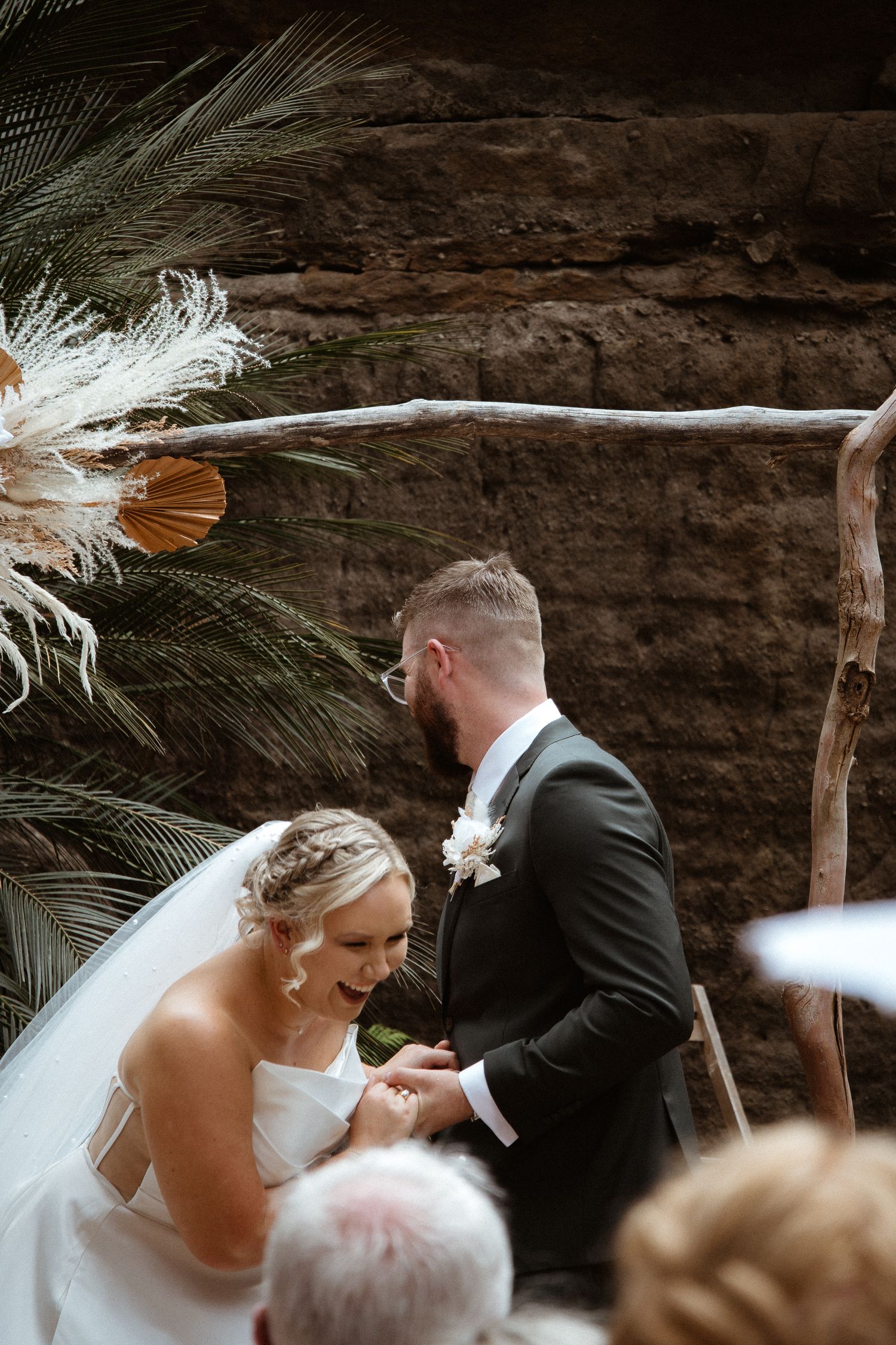 Intimate wedding moment captured under rustic wooden arch with pampas grass and palm decor.