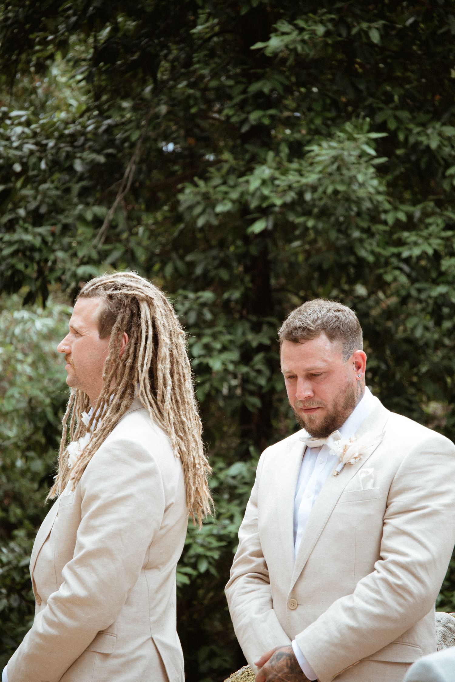 Two people in cream colored suits stand back-to-back during wedding ceremony outdoors.
