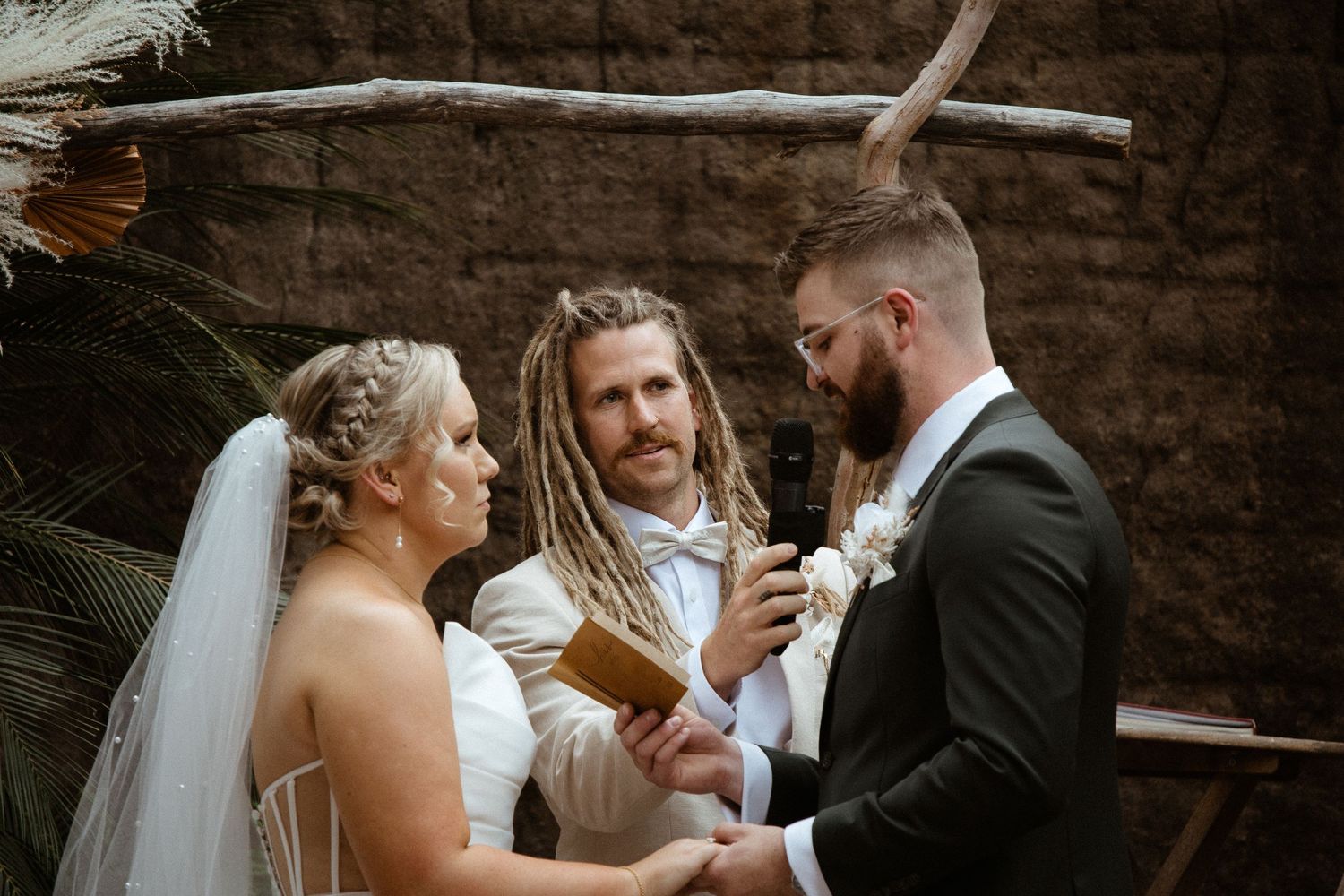 Wedding ceremony with rustic wooden arch and officiant in white suit against stone wall.