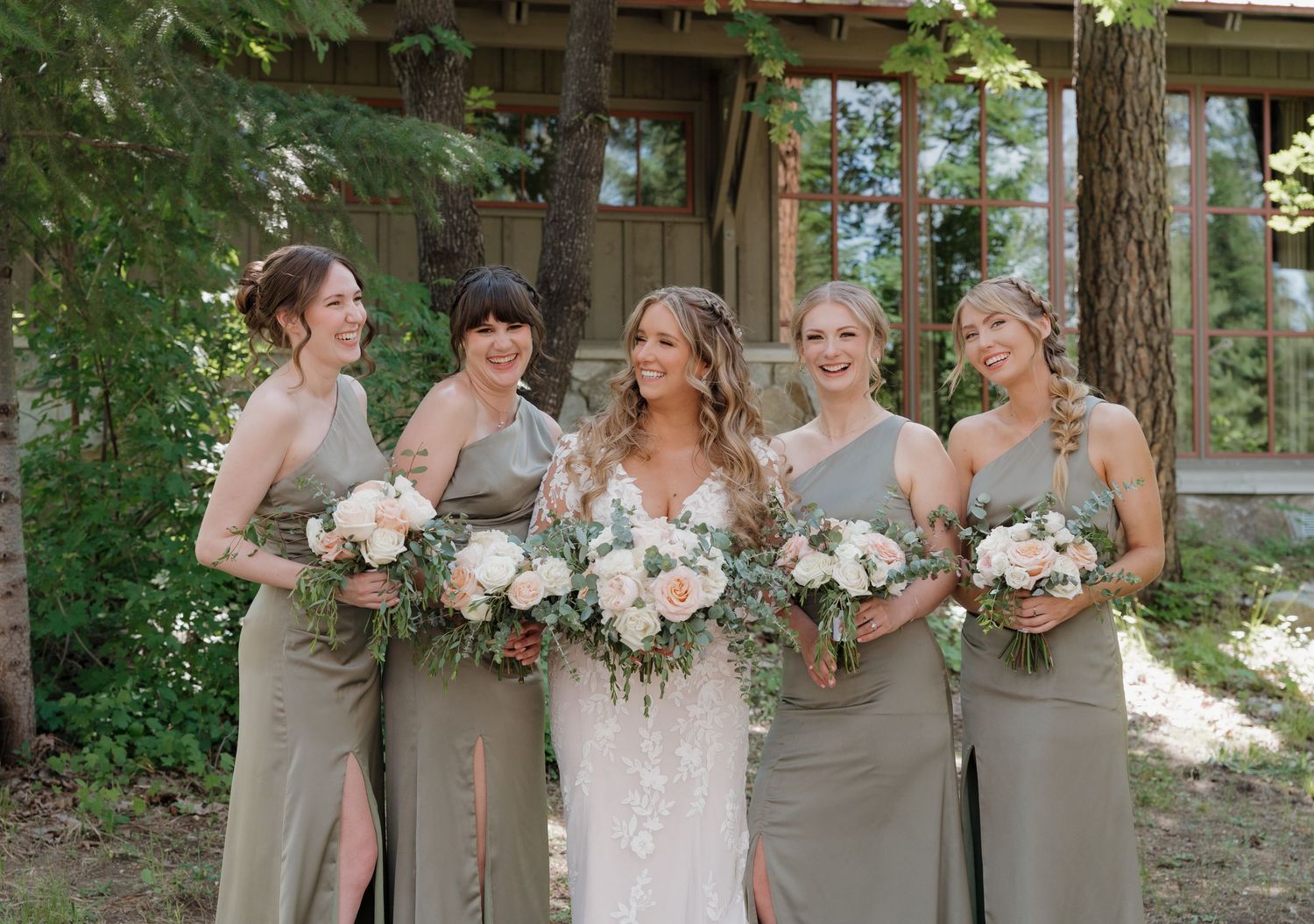 Bridesmaids in elegant taupe dresses hold white and green bouquets while posing together outdoors at a rustic venue.