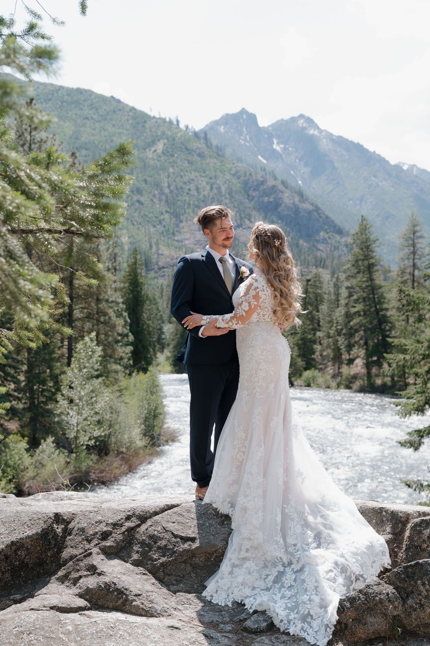 Couple in wedding attire embrace on rocky outcrop with snow-capped mountains and evergreen forest in background.