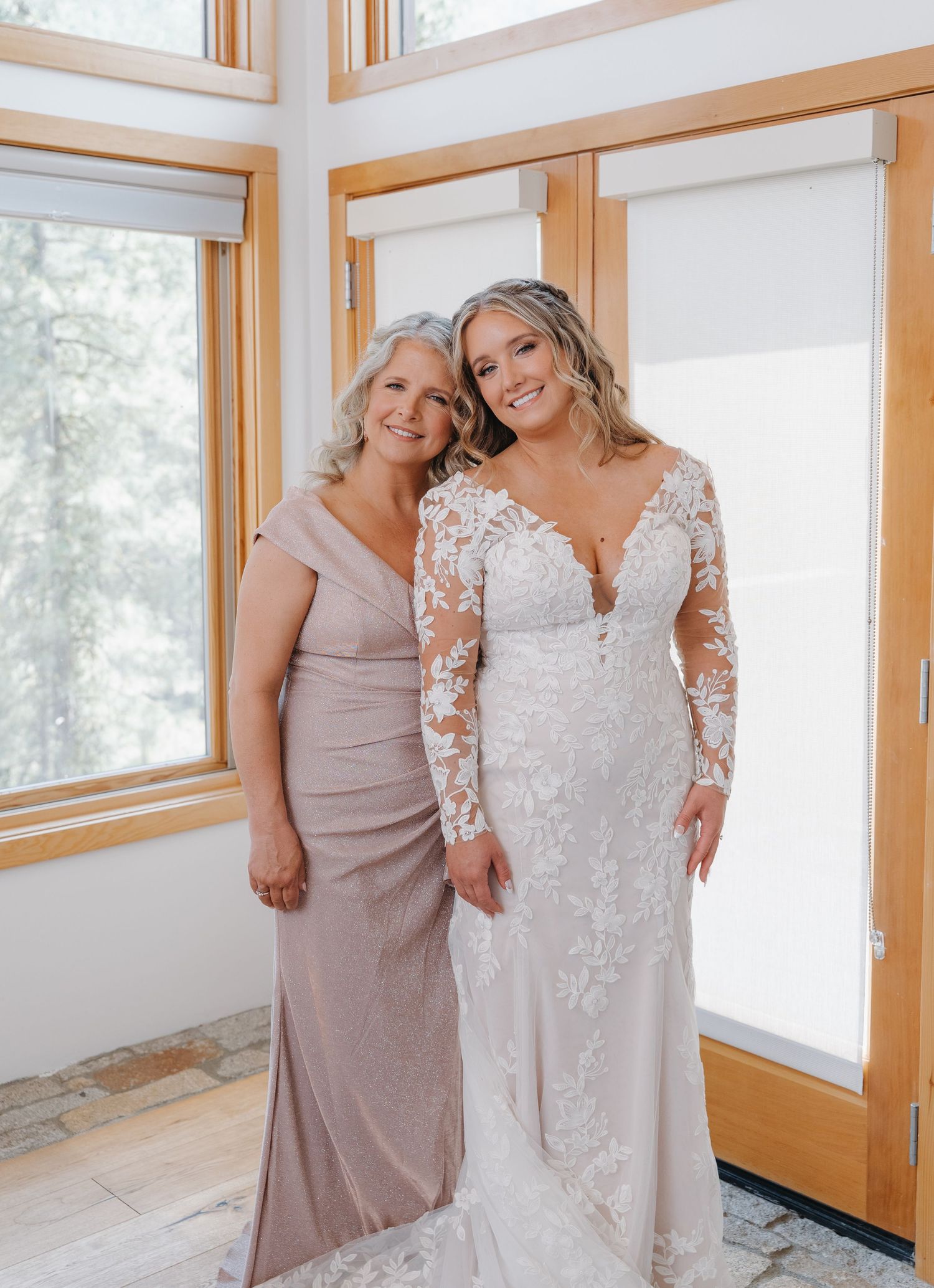 Two people in a sun-filled room share a tender moment, one wearing a taupe dress and one in an elegant lace wedding gown.