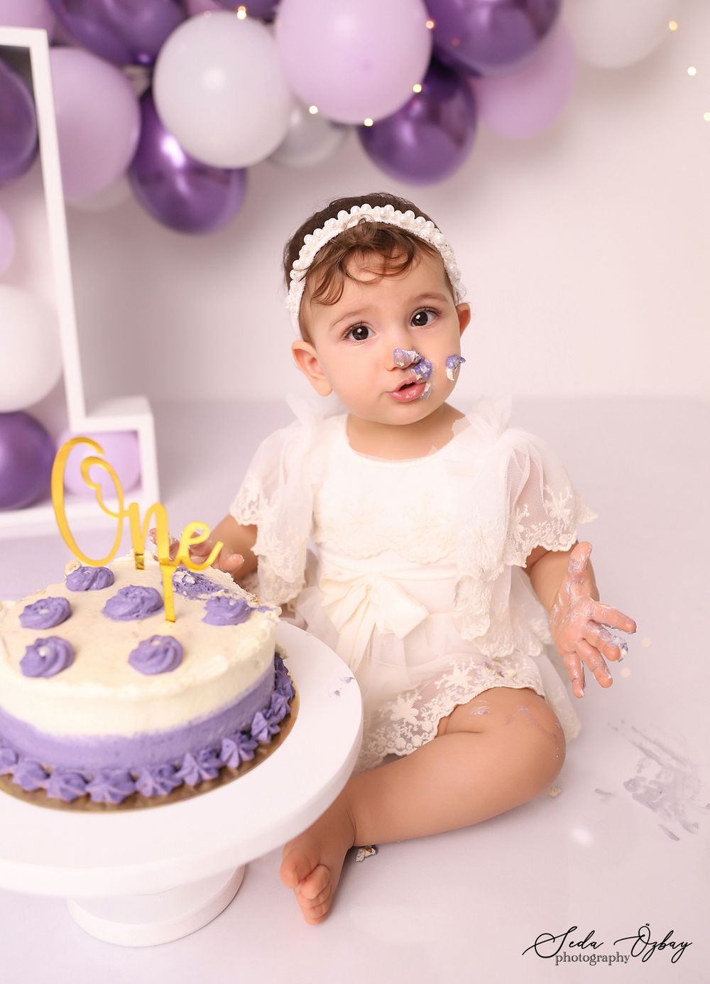 Cake smash photo session with purple decorations and a white birthday cake.