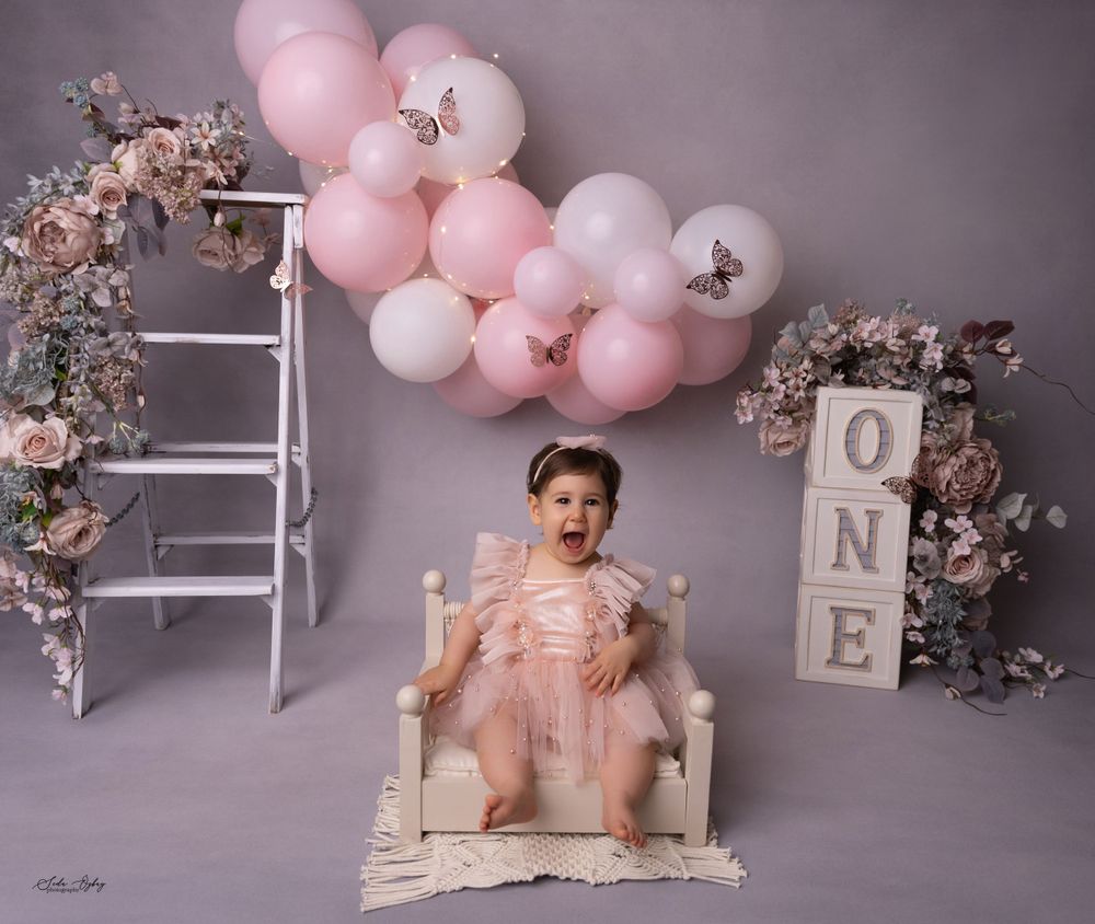 First birthday setup with pink balloons, floral decor, and white ladder against grey backdrop.