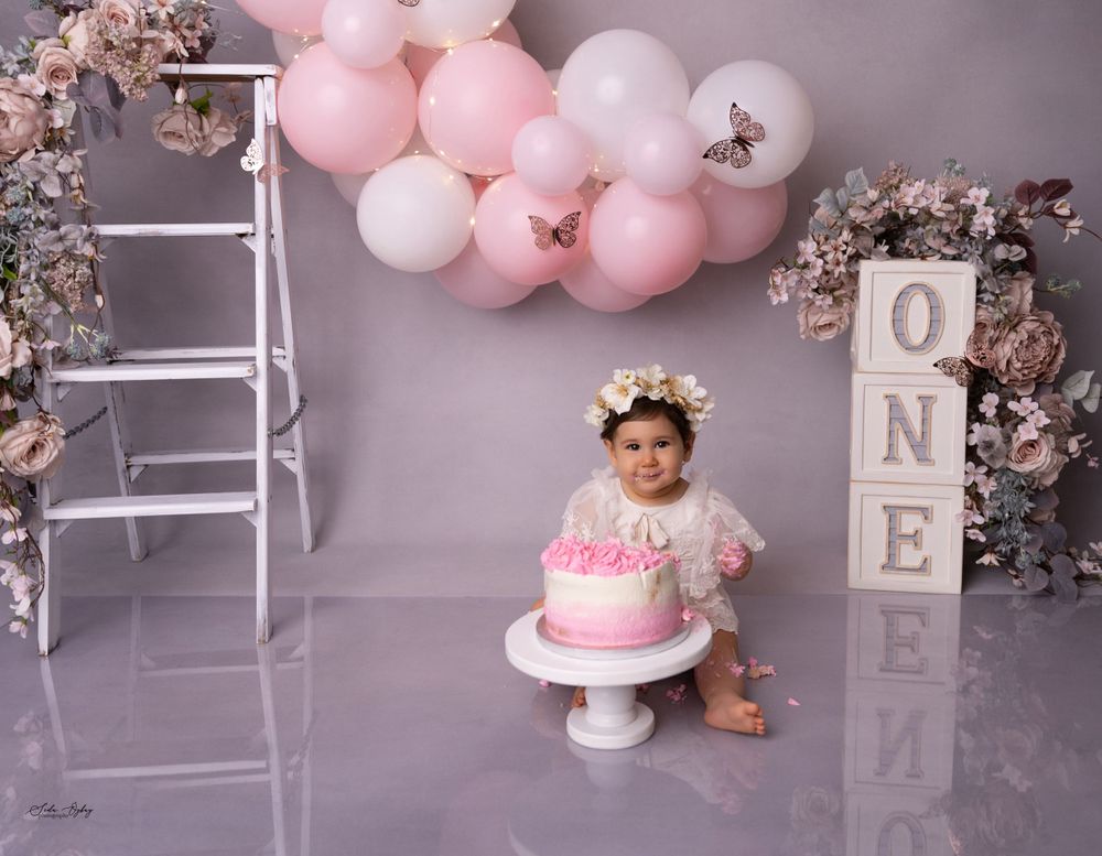 Baby with birthday cake in pink and white themed setup with balloon arch and floral details.