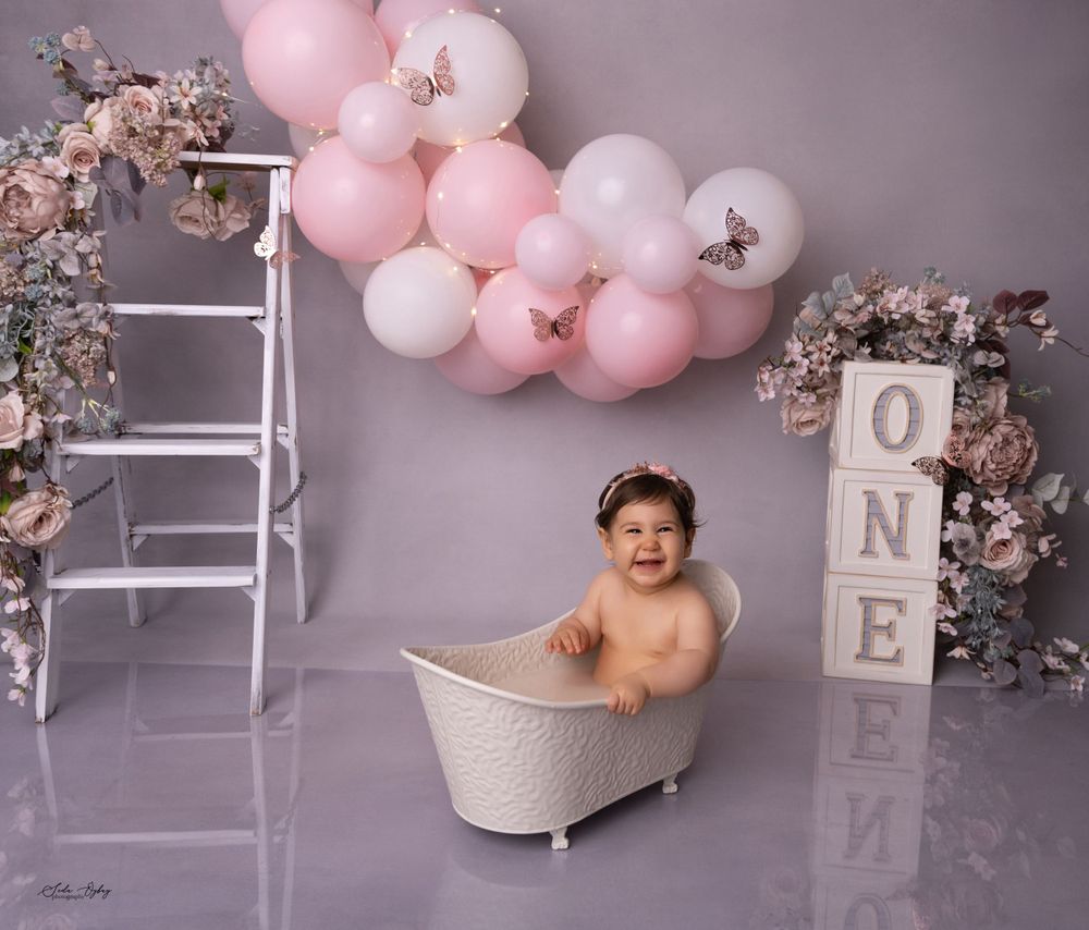 Baby in white bathtub with pink balloon arch and floral first birthday decorations.