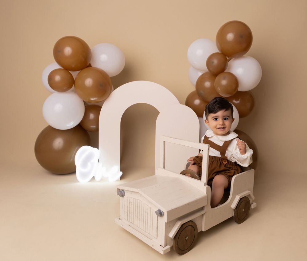 Baby sitting in vintage white toy car with brown and white balloon arch backdrop.
