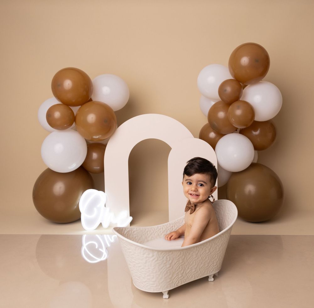 Baby sitting in white decorative bathtub with balloon arch and neutral backdrop.