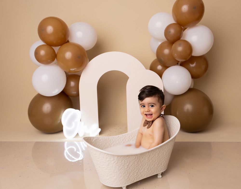 Baby posing in white miniature bathtub against brown and white balloon display.