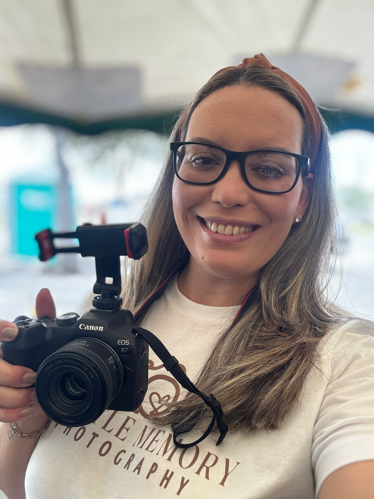 Smiling female photographer holding a Canon EOS camera at an outdoor event wearing glasses.