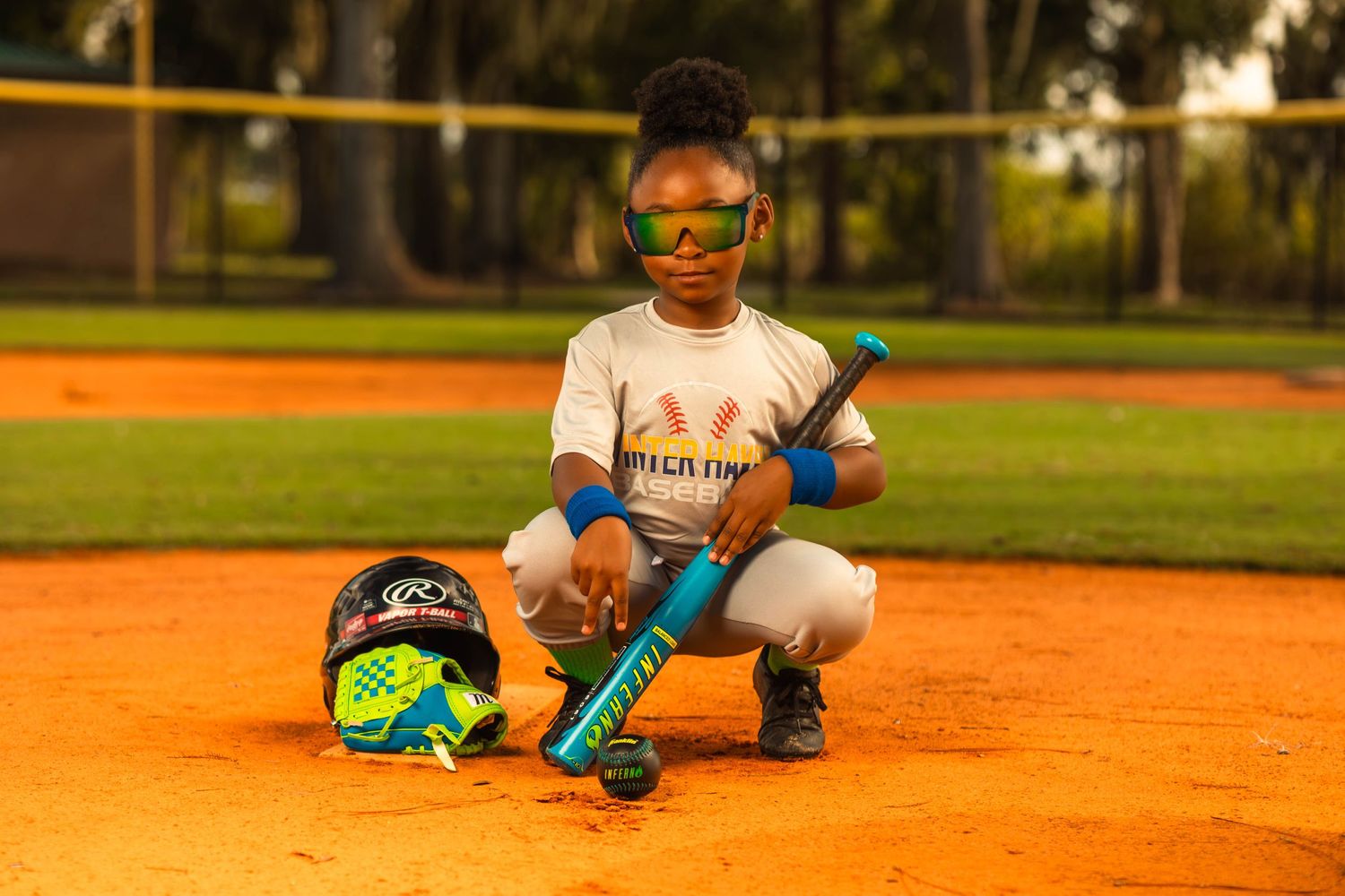 Young girl softball player crouches on dirt field with bat, helmet, and glove beside her.