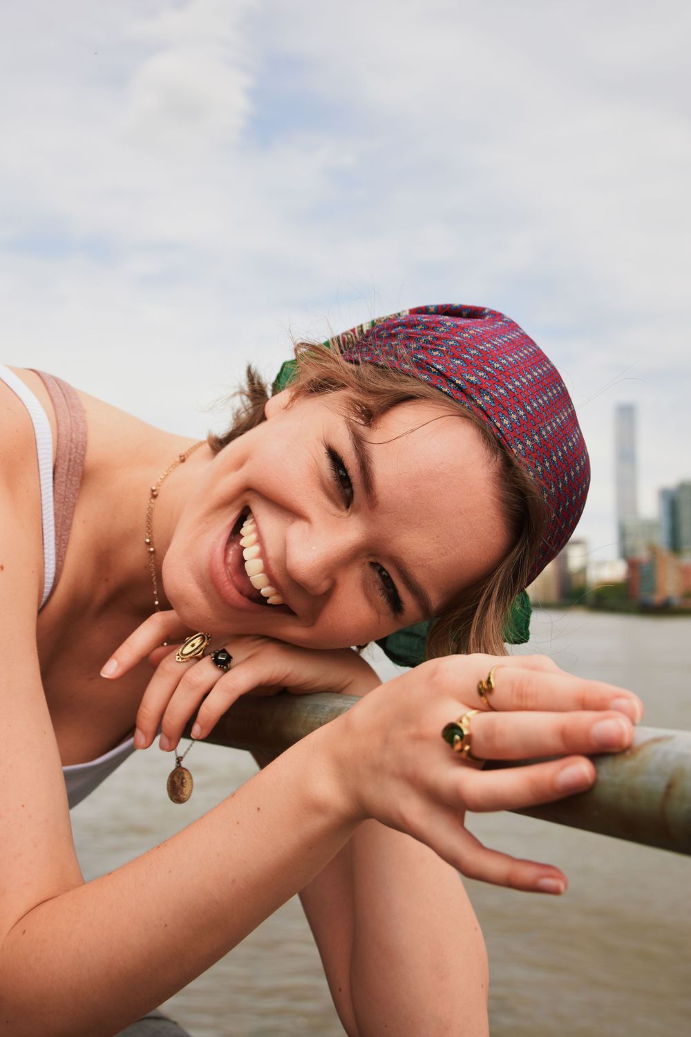 Young person wearing a patterned cap smiling joyfully by waterfront with rings and jewelry.
