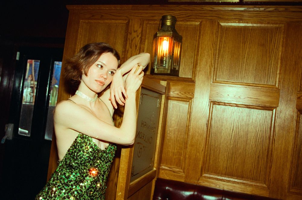 Woman in green sequin dress posing by wooden wall with vintage lantern in dimly lit bar.