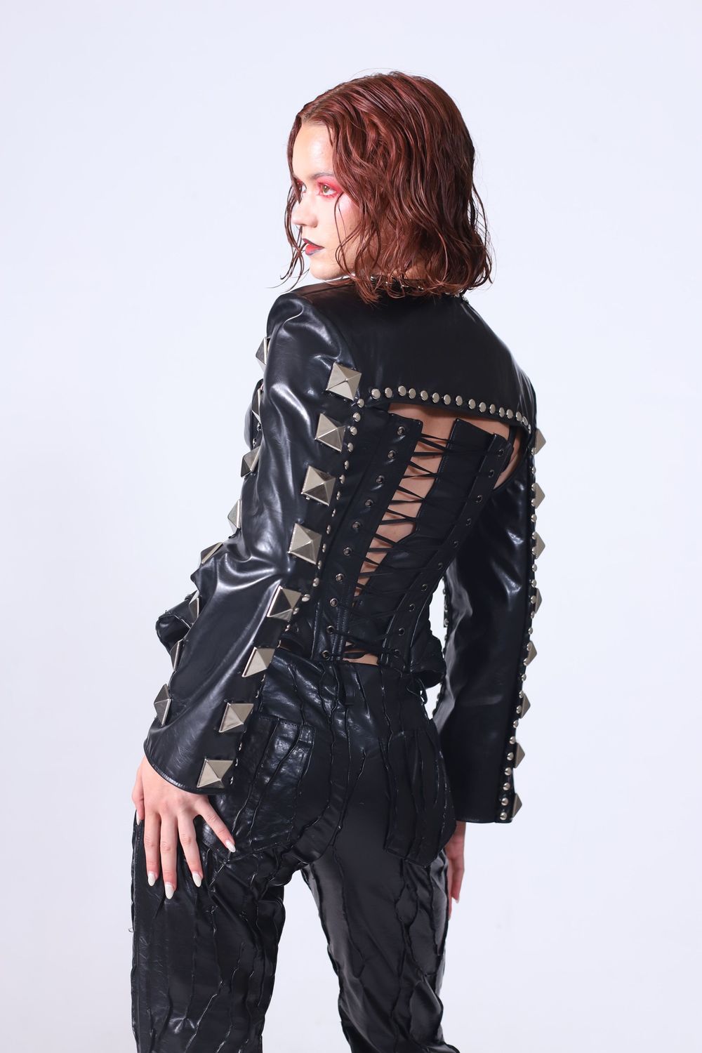 Woman in black studded leather corset jacket with lace-up back detail, paired with black pants, posing against white background.