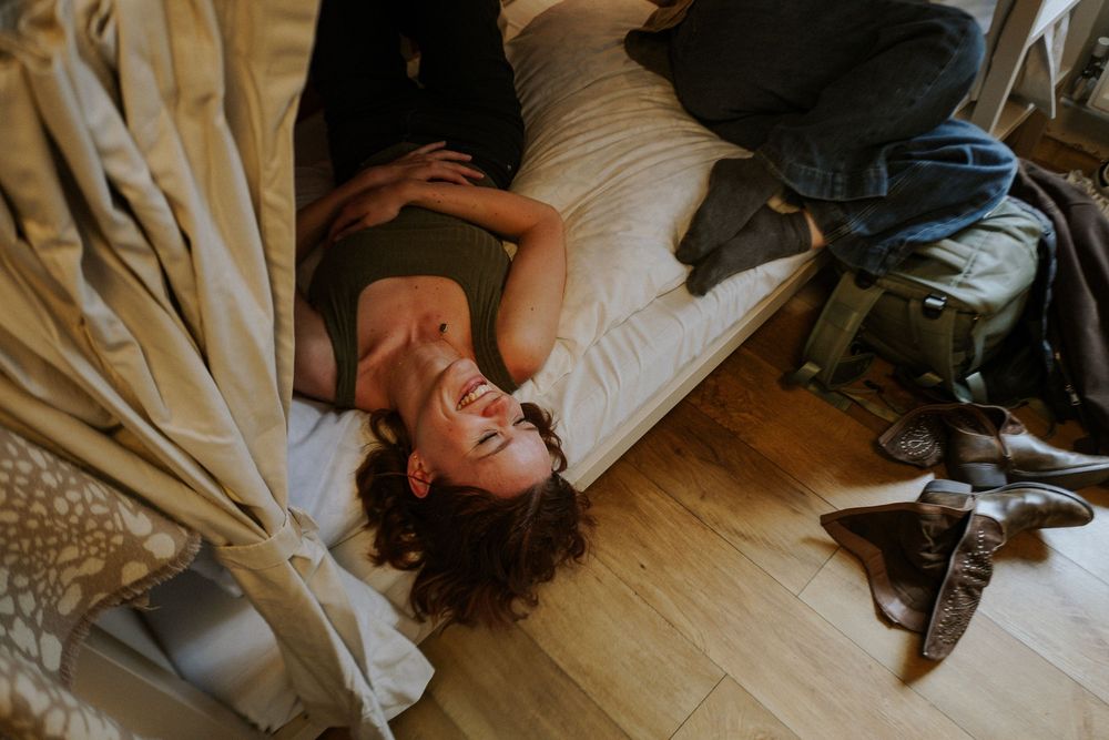 Woman lying on bed laughing, with backpack and boots on wooden floor nearby.