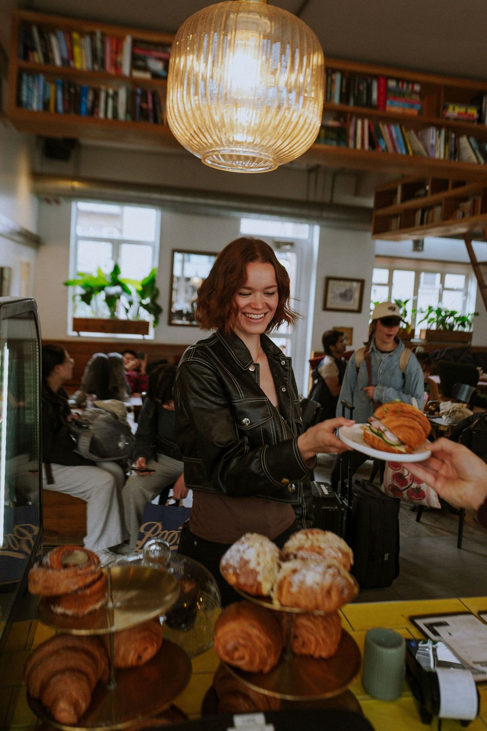 Smiling barista in leather jacket serves a croissant sandwich at a cozy cafe counter with pastries and books.