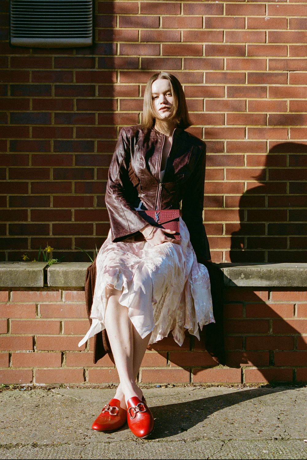 Figure in vintage-inspired outfit with brown leather jacket, flowy white skirt and bright red shoes sits on brick steps.
