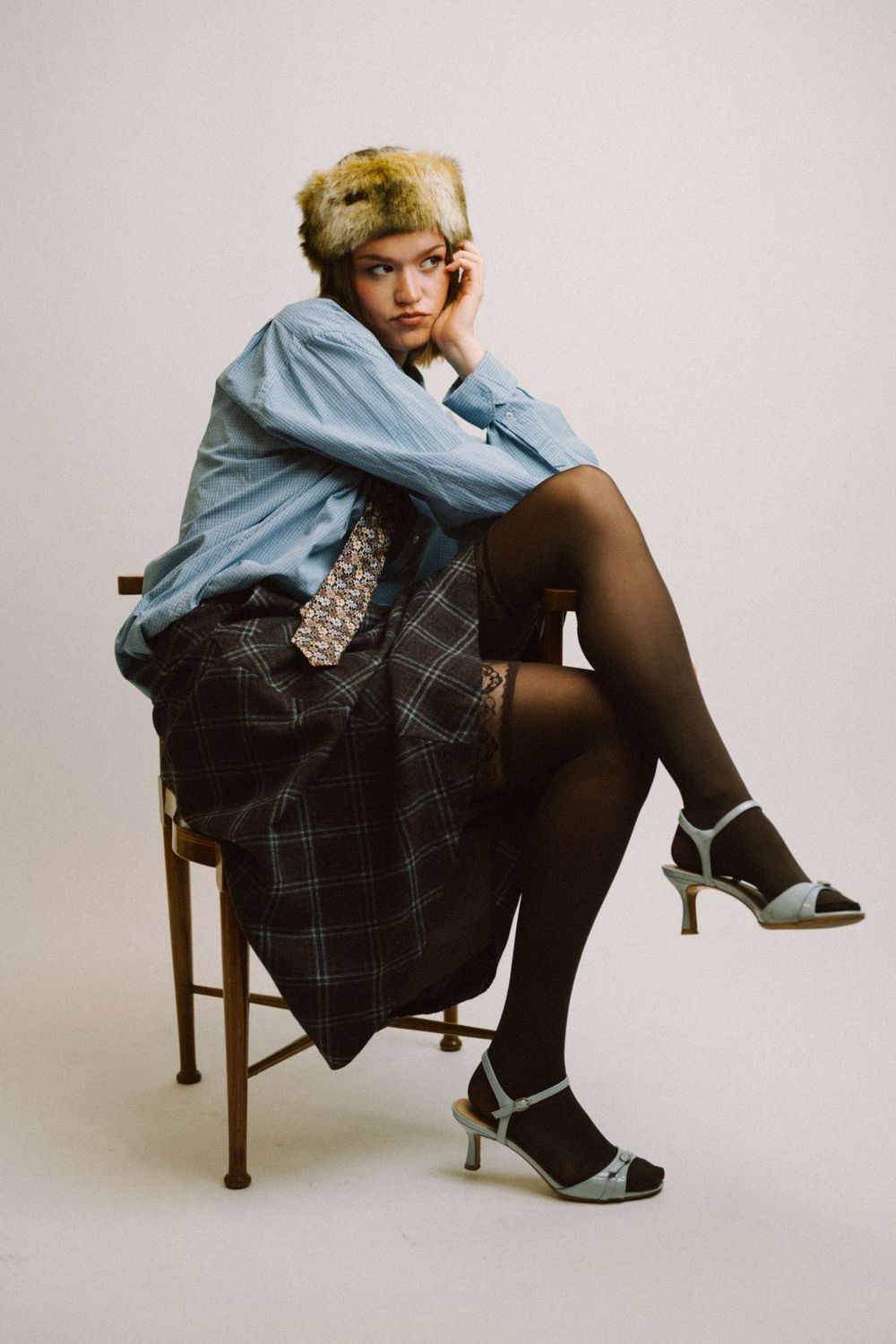 Fashion model in light blue top and plaid skirt poses thoughtfully on wooden chair in studio setting.