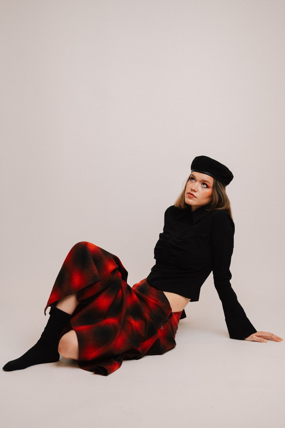 Fashion model in black beret and red plaid dress poses elegantly on white studio floor in artistic photo series.