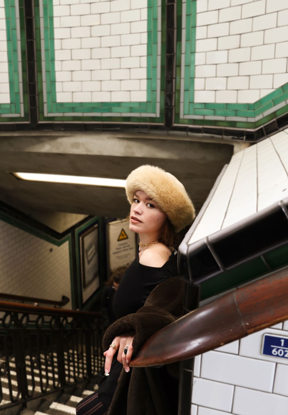 Young woman in a tan beret and black coat poses on vintage London Underground station stairs.