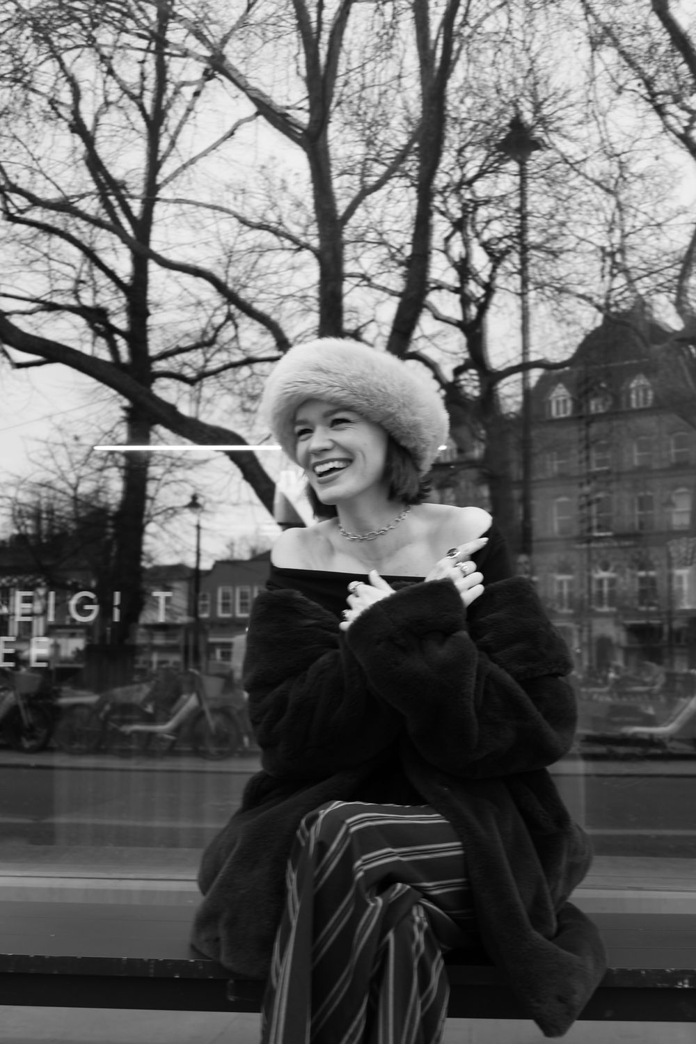 Black and white photo of a smiling young woman in fur hat and coat sitting outdoors in winter.