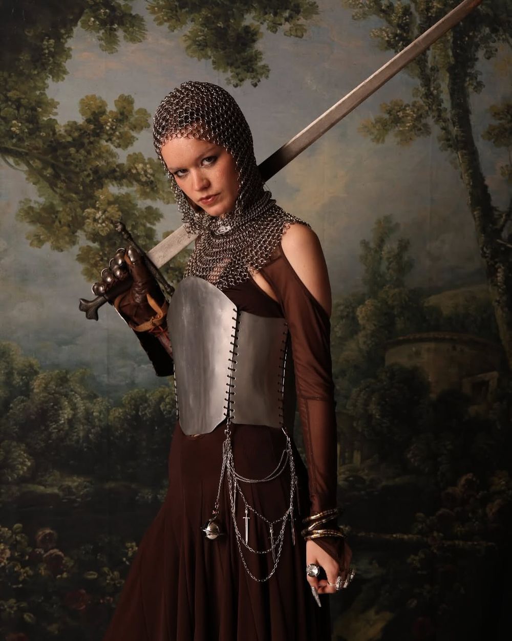 Medieval-inspired portrait with chainmail coif, armor breastplate, and long sword against a pastoral landscape background.