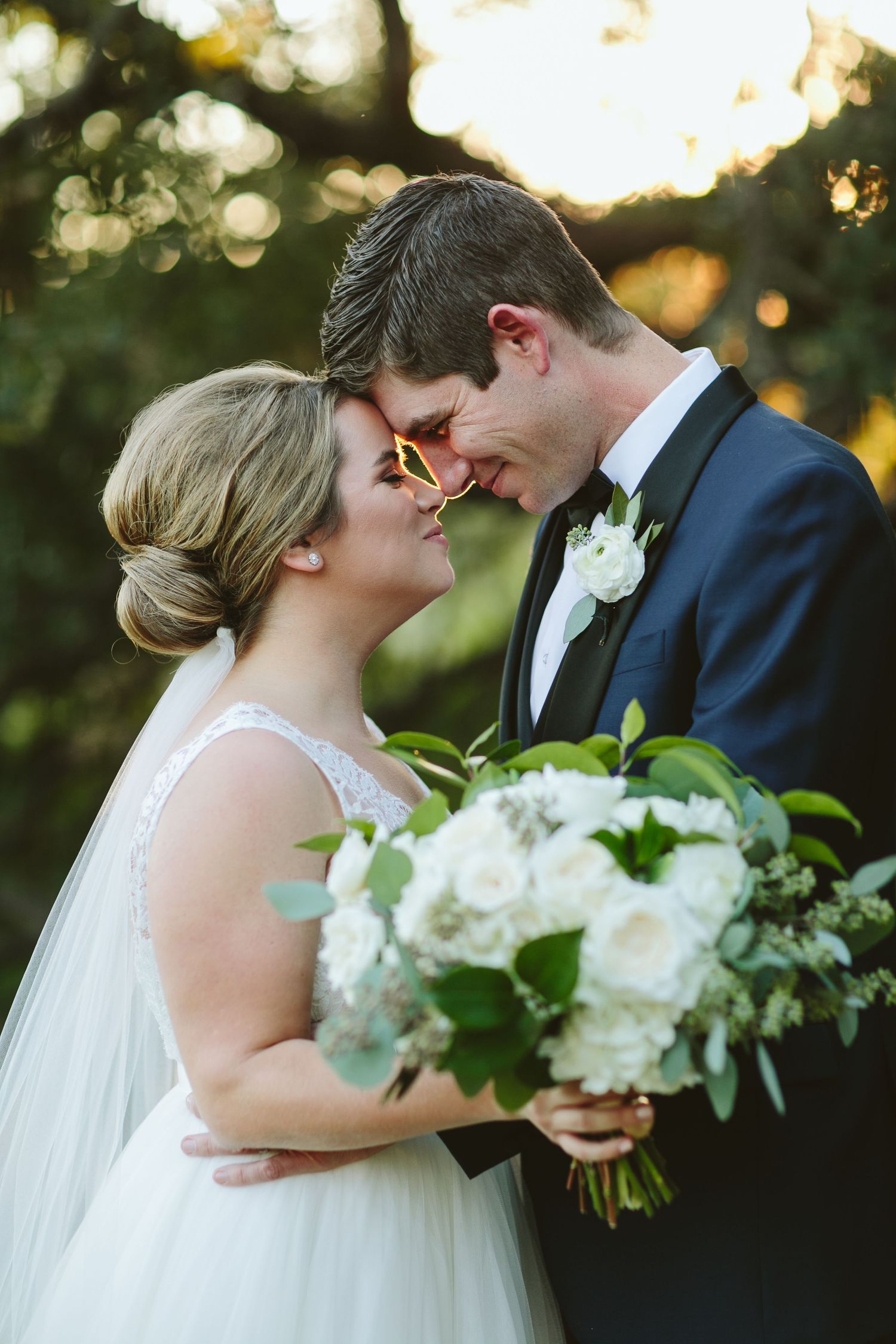 Bride and groom share intimate moment in garden setting with white floral bouquet.