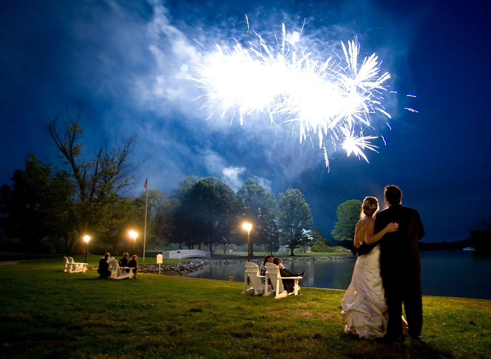Silhouettes watch fireworks illuminate the night sky over a lake while celebrating outdoors.