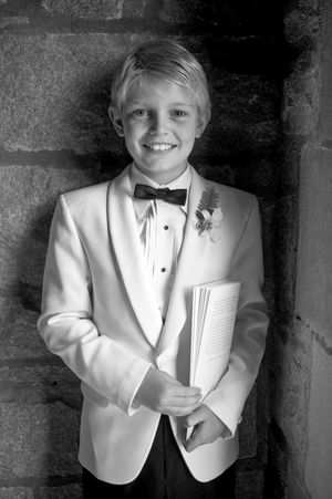 A young attendant in a white tuxedo jacket and bow tie holds a program at a formal event.