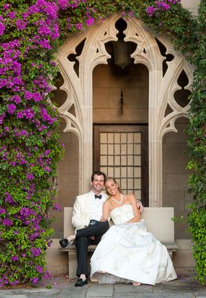 A romantic wedding couple relaxes in an elegant stone archway adorned with vibrant purple bougainvillea flowers.
