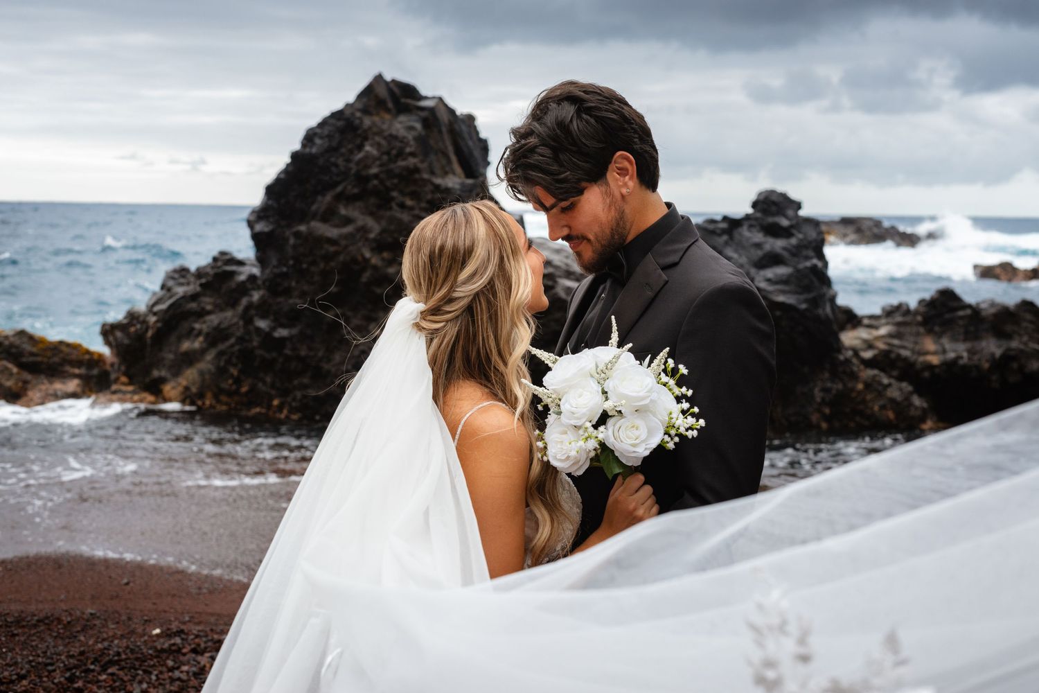 Bride holding white bouquet with flowing veil embraces groom near black lava rocks and crashing ocean waves.