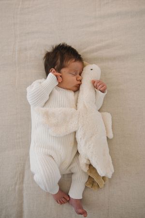 A sleeping newborn in a white knit outfit lies on beige linen bedding with a soft white plush toy.