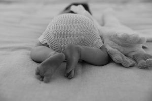 A sleeping infant in a knit sweater lies on their stomach on a soft white blanket, photographed in black and white.