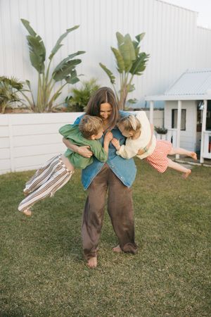 A person in casual clothing plays with two young children on a grassy lawn with tropical plants in the background.