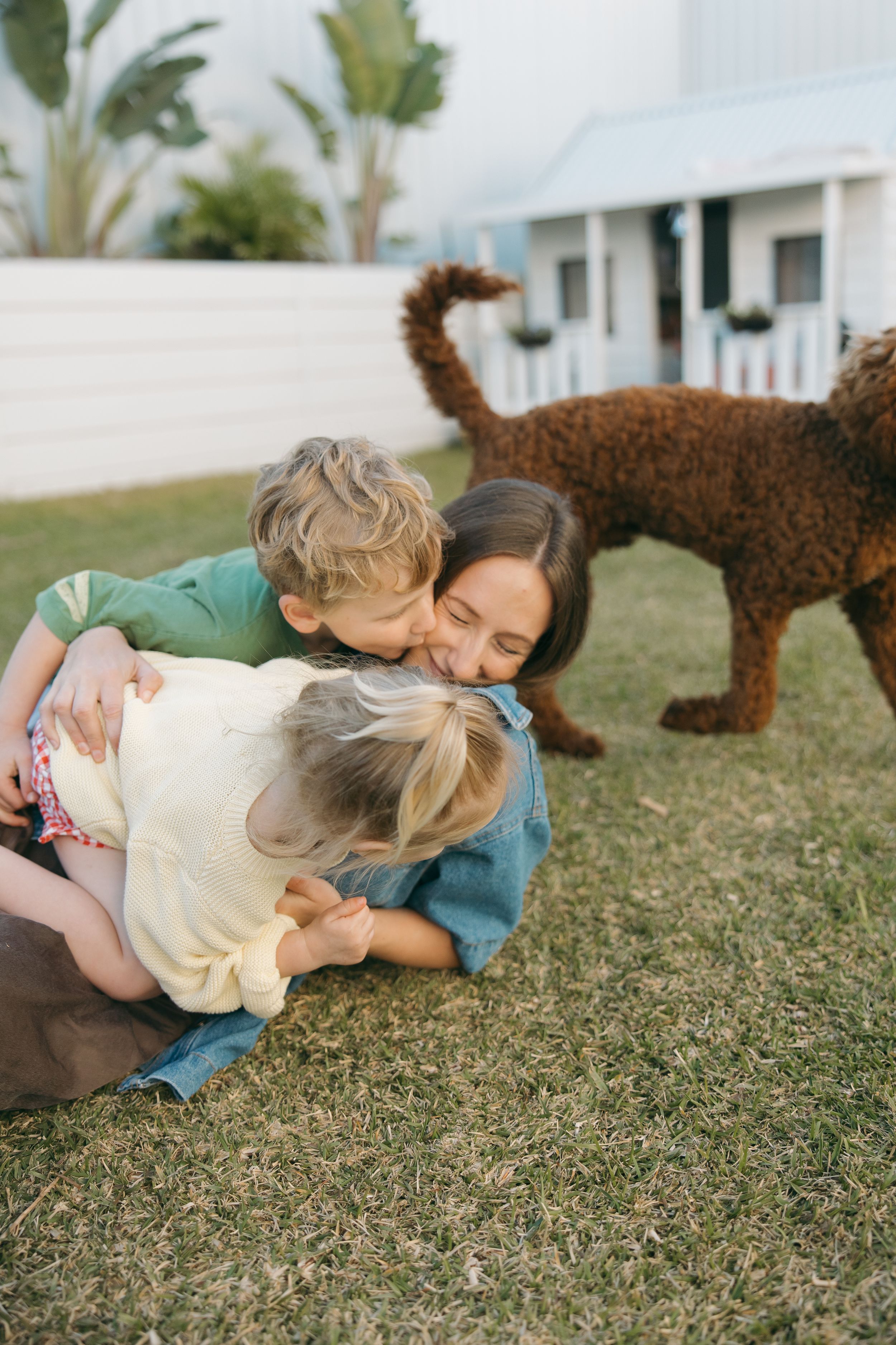 Two young people lay on grass while a brown dog walks near them in an outdoor setting.