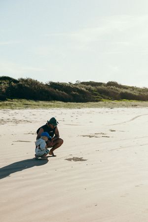 Figure crouches on a pristine beach with sand dunes and vegetation in the background.